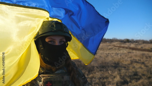 Female military medic of ukrainian army looks into camera against waving blue-yellow banner. Portrait of girl in camouflage uniform and helmet holding flag of Ukraine at field on sunny day. Dolly shot