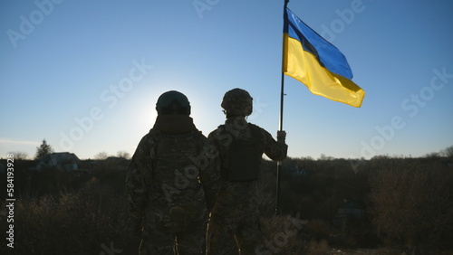Female and male soldier of ukrainian army standing at peak of hill with raised flag of Ukraine. Military couple in camouflage uniform holds hands of each other looking at blue sky with sun. Dolly shot