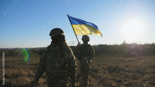 Young woman and man in military uniform going with national banner at meadow. Female and male soldier of ukrainian army walking with flag of Ukraine at field. Victory against russian aggression