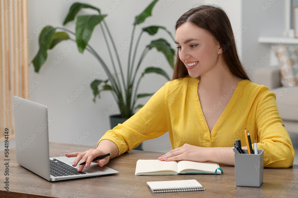 Happy young woman with notebook working on laptop at wooden table indoors Happy young woman with notebook working on laptop at wooden table indoors