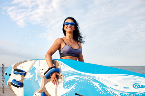 Woman 40-45 years old, walking along the beach against the background of water and beach umbrellas with a surfboard, going to ride the waves, S3niorLife, waiting for a wave, surfing as a lifestyle © Georgii