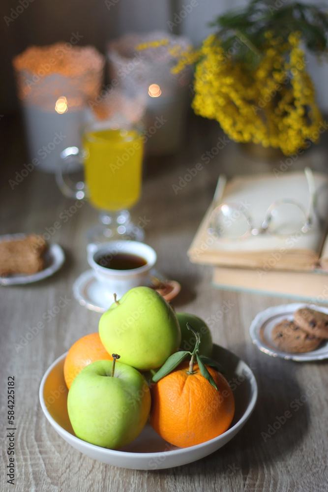 Cup of tea, plates with cookies, glass of orange juice, books, reading glasses, bowl of fruit and candles on the table. Selective focus. Cup of tea, plates with cookies, glass of orange juice, books, reading glasses, bowl of fruit and candles on the table. Selective focus.