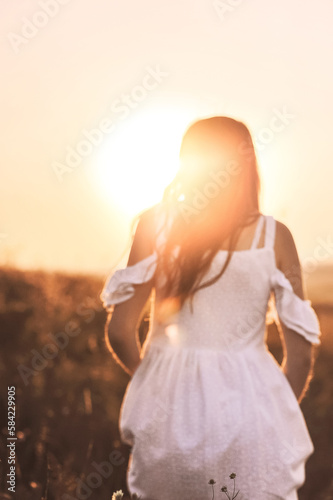 Rear view of young woman in summer white dress walking on sunset background. Attractive girl looking into the distance at nature