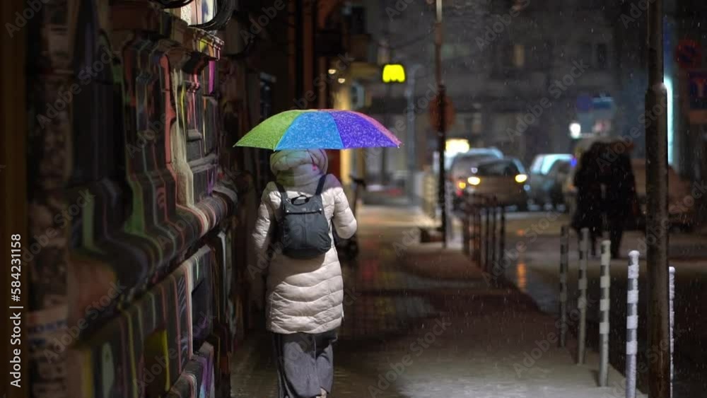 Woman in white coat holds rainbow umbrella, walks under street light at night Woman in white coat holds rainbow umbrella, walks under street light at night