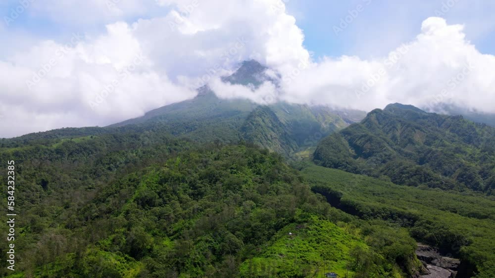 Aerial view of slope of mountain overgrown with forest and meadow which is split by valley as lava flows when a volcano erupts. Seen peak of the mountain shrouded by clouds - Merapi Volcano, Indonesia Aerial view of slope of mountain overgrown with forest and meadow which is split by valley as lava flows when a volcano erupts. Seen peak of the mountain shrouded by clouds - Merapi Volcano, Indonesia