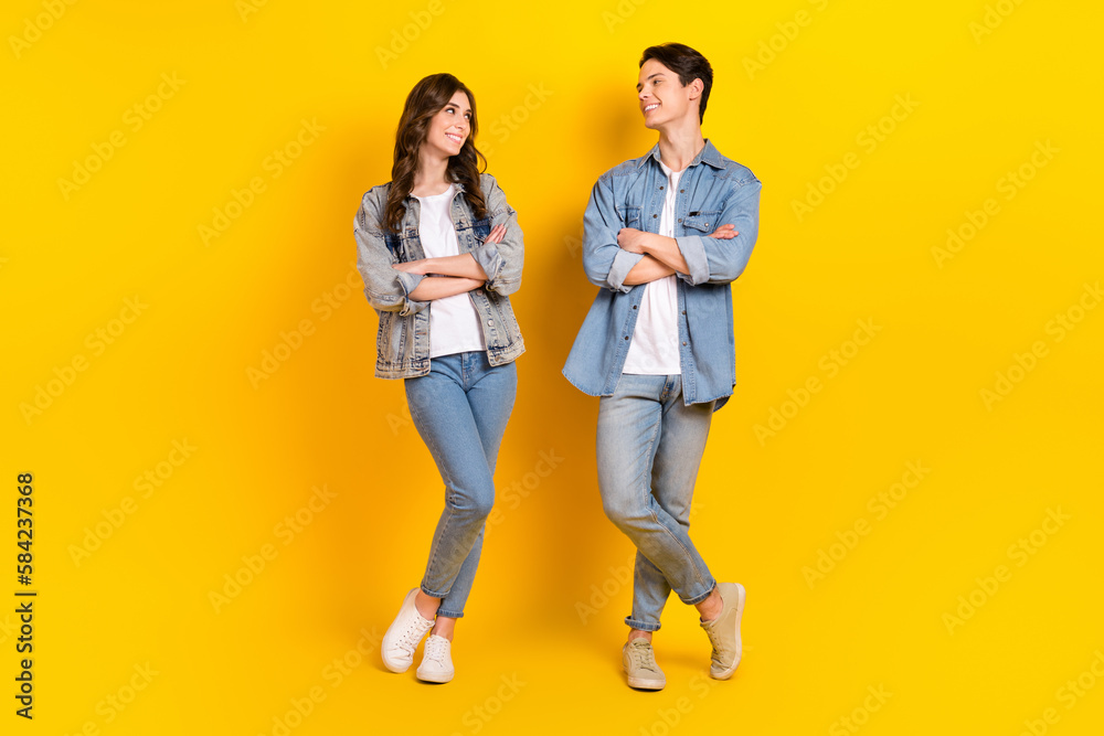 Full length portrait of two cheerful satisfied people crossed arms look each other isolated on yellow color background Full length portrait of two cheerful satisfied people crossed arms look each other isolated on yellow color background