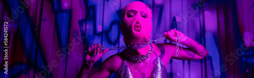passionate woman in balaclava posing with silver chain near graffiti in purple light, banner.