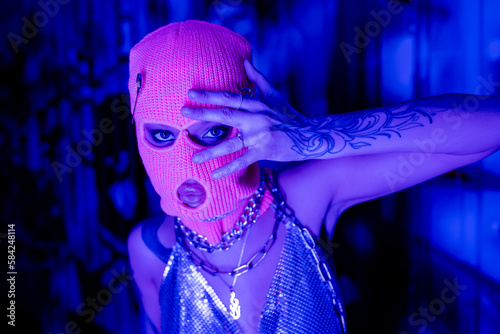 provocative woman in balaclava and metallic top with necklaces posing with hand near face in blue and purple lighting. photo