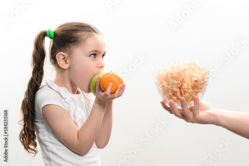 Big bowl of chips and fruit, beautiful little girl eats an apple, child is given a bowl of chips, white background and copy space.