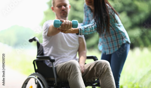 Female physiotherapist helps disabled man in wheelchair to lift weight of arm in park © H_Ko