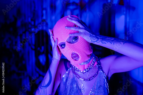passionate tattooed woman in silver neck chains looking at camera while touching knitted balaclava in blue and purple light. photo
