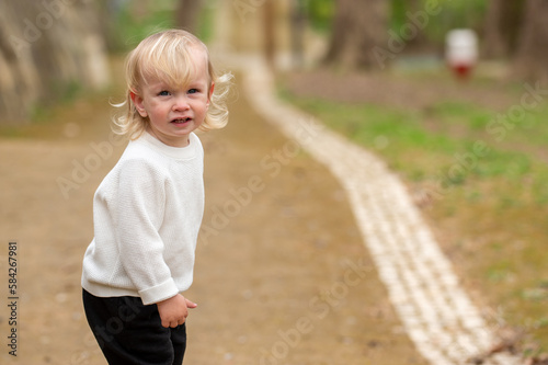 Cute blonde kid in a white sweater is walking around a park. Interested caucasian baby boy alone on a sidewalk on a chilly day. A boy in a white sweater exploring the world outdoors.