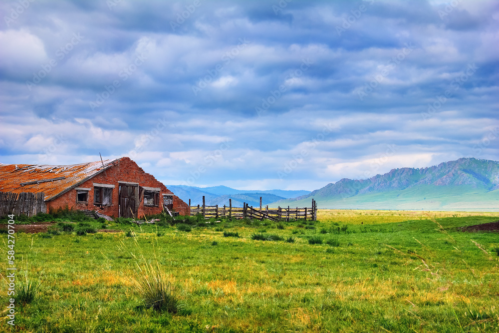 Beautiful scenery with old abandoned farm. Ruined building with a wooden fence and green field at the background of mountain range and cloudy sky in Khakassia, Southerh Siberia, Russia