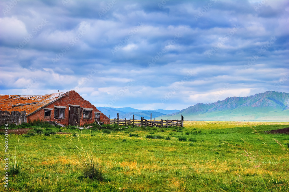 Obraz premium Beautiful scenery with old abandoned farm. Ruined building with a wooden fence and green field at the background of mountain range and cloudy sky in Khakassia, Southerh Siberia, Russia