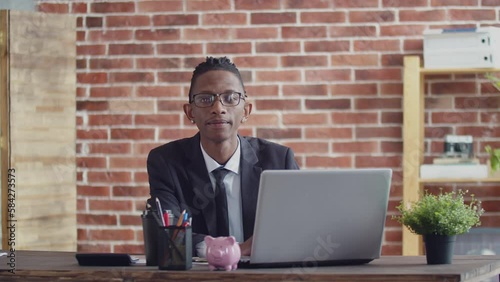Black employee with glasses and a business suit sits in a cozy office against the backdrop of a brick wall, works at a notebook, nods knowingly and looks at the camera photo
