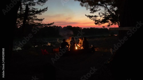 Group of young people are sitting on logs around fire on the banks of river at sunset roasting sausages on sticks. Slow motion