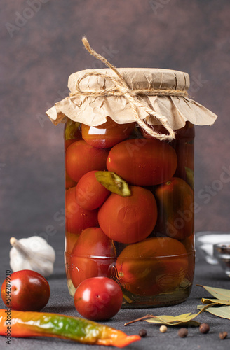 Homemade pickled tomatoes with chili peppers and parsley in glass jar on brown background, vertical image