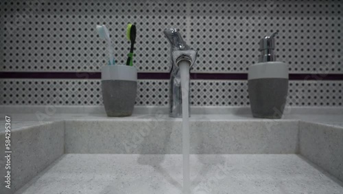 Water flows from tap to washbasin in bathroom closeup. Woman hands close lever of tap. Toothbrush and soap with dispencer located near tap photo