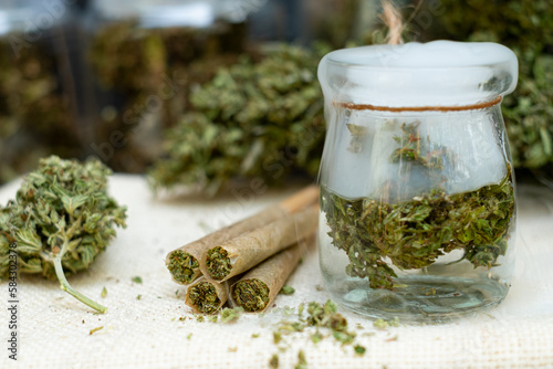 Marijuana joint pre-rolled cone paper with cannabis buds in a clear glass jar sit on a smoky floor