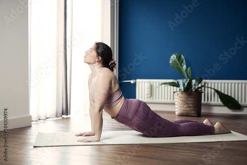 Young sporty woman practicing yoga at home, doing upward facing dog exercise, Urdhva mukha shvanasana pose, working out, wearing sportswear, pants and top, indoor full length, Yoga in living room photo