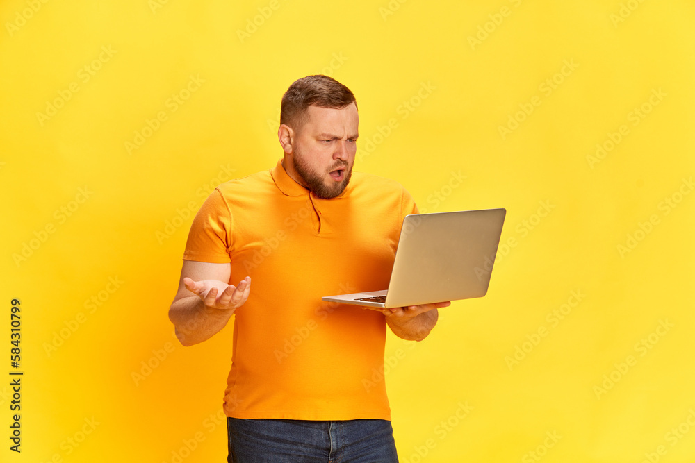 Disappointed sales man trying to understand what happening, holding laptop with questioning facial expression over yellow background