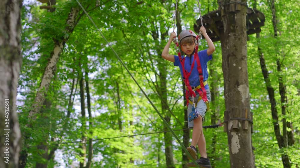 A little boy is enjoying an exciting adventure on a rope obstacle course in a lush forest. He is wearing a helmet and a safety harness as he navigates through the challenging course, showing bravery
