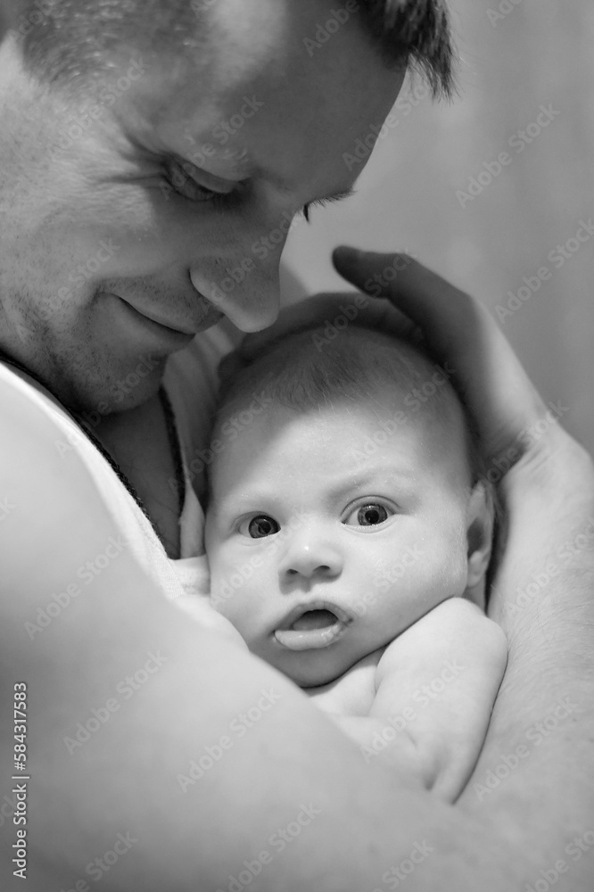 Happy father gently holds a newborn baby in his arms. The child looks into the lens. Father's day concept. Black and white photography. Lifestyle documentary moment. Vertical photo. Happy father gently holds a newborn baby in his arms. The child looks into the lens. Father's day concept. Black and white photography. Lifestyle documentary moment. Vertical photo.