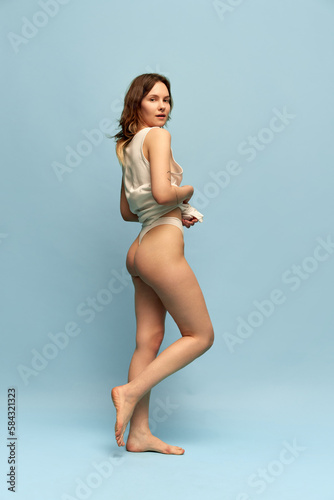 Full-length image of slim, beautiful, young girl in comfortable homewear posing against blue studio background. Anti-cellulite care. Concept of body and skin care, figure, fitness, health, wellness.