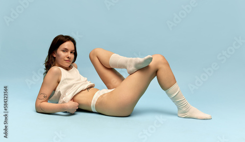 Portrait of beautiful young girl with slim body, in comfortable homewear and socks posing against blue studio background. Coziness. Concept of body and skin care, figure, fitness, health, wellness.