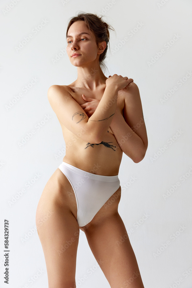 Portrait of young slim girl with tattoos, covering breast with hands, posing in underwear against grey studio background. Concept of body and skin care, figure, fitness, health, wellness. Portrait of young slim girl with tattoos, covering breast with hands, posing in underwear against grey studio background. Concept of body and skin care, figure, fitness, health, wellness.