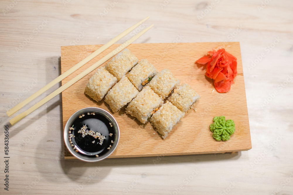 Fototapeta premium Traditional Japanese food. Freshly made sushi rolls of fish, rice, cheese, cucumber, sesame seeds served on a wooden board. Next to it: soy sauce, ginger, wasabi, chopsticks. 