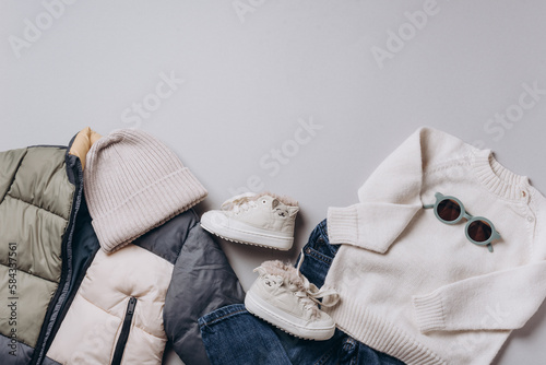 Fashion children's clothing, shoes - colorblock puffer jacket, knitted hat, scarf, boots, jeans, sunglasses on grey background. Spring, autumn collection. Top view, flat lay. Beige clothes outfit.