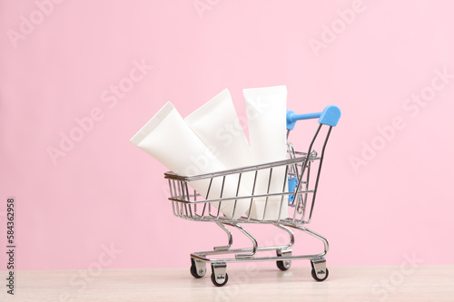 Mini Shopping cart with white cream tubes on pink background. Beauty concept