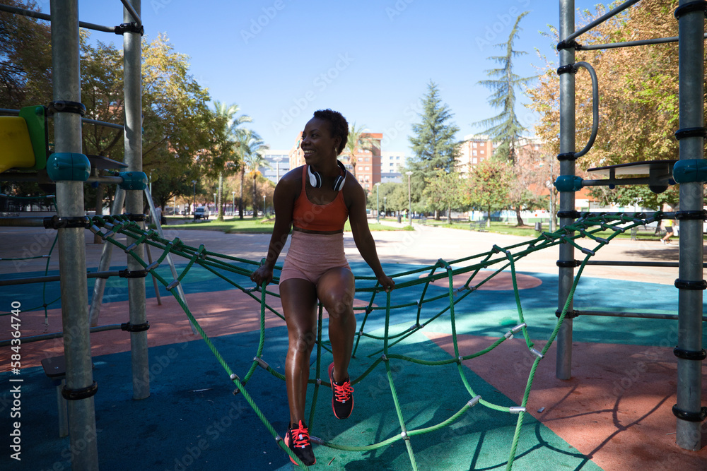 Fototapeta premium Young and beautiful Afro American woman with sculpted body sitting in the park before training. She is dressed in an orange top and tights and listens to music with white headphones. Sport and health.