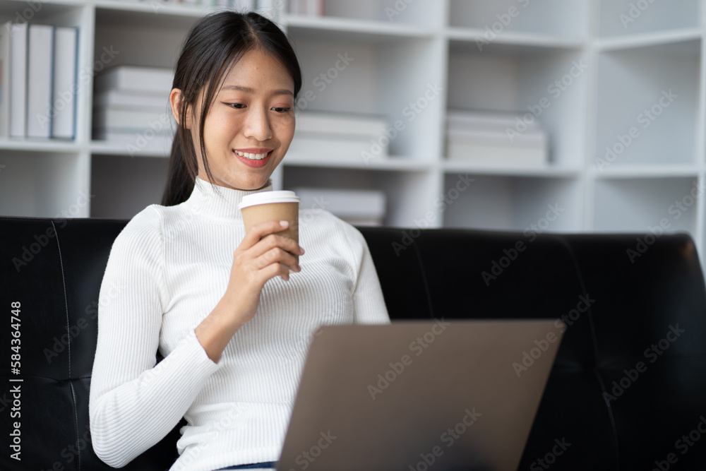 Happy young Asian girl using laptop computer while holding a coffee on the sofa at home. Happy young Asian girl using laptop computer while holding a coffee on the sofa at home.