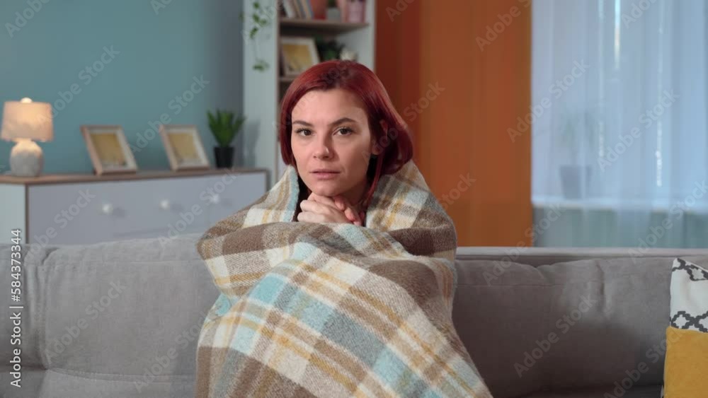 lack of heating due to debts or breakdown of heating systems, portrait of a sad woman warms hands wrapped in warm blanket while sitting on sofa looking at camera