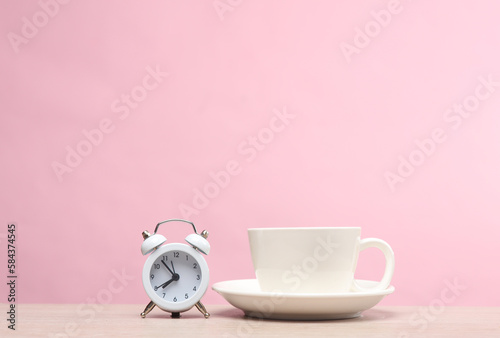 Ceramic white coffee cup with alarm clock on the table, pink background