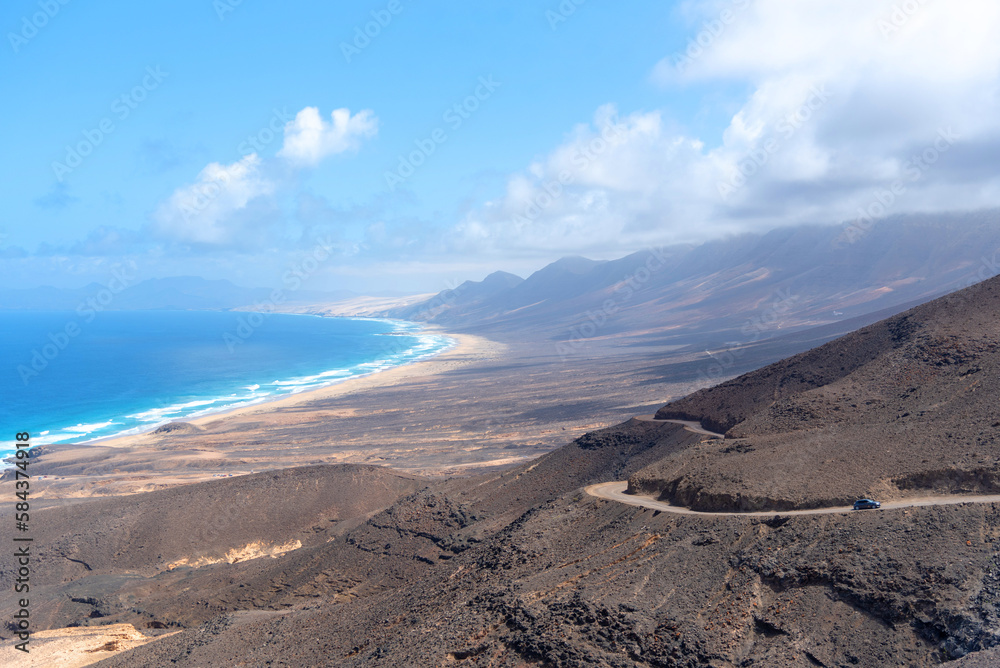 Vista panorámica de la playa de Cofete con agua turquesa, arena blanca y algunas olas, rodeada de un paisaje volcánico y desértico durante un día soleado con cielo azul en Fuerteventura, Isla Canaria
