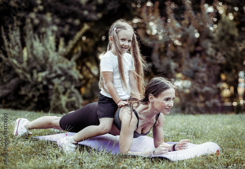 Athletic woman mom with daughter on her back is practicing plank exercise in the park. They spend time together. Healthy family