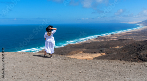 joven rubia de pelo largo vestida de blanco sosteniendo un sombrero de paja de espaldas disfrutando de la playa de arena blanca y aguas cristalinas de Cofete en Fuerteventura Islas Canarias. photo