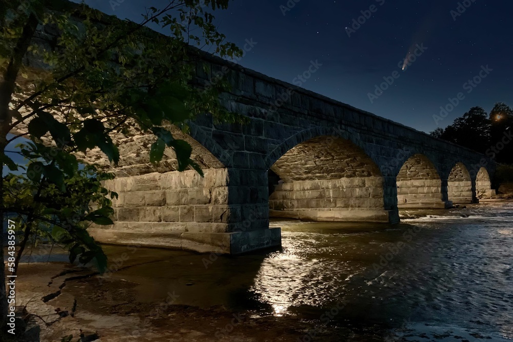 Fototapeta premium Low angle view of a stone bridge with a Comet visible passing in the sky during the night