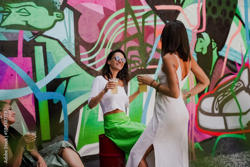 Three stylish young women dancing and laughing with drinks in hand against a colorful graffiti wall. A lively urban summer vibe full of joy, friendship, street art, and carefree energy.