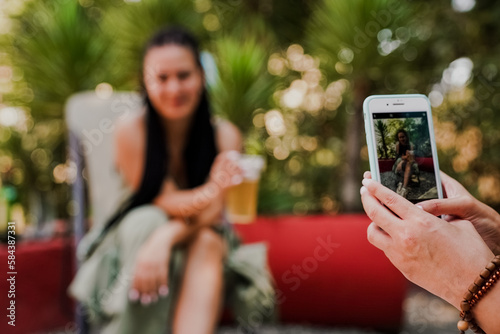 Close-up of a hand taking a photo of a smiling woman with a drink using a smartphone. Summer outdoor scene with palm trees, relaxation, and casual social moments captured digitally.