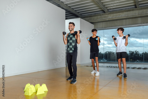 Total Body fitness group of teenagers training in dumbbell fitness class. Full length of male coach instructor and Two boys performing exercise with dumbbells in a big gym with windows. Copy-space