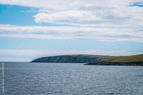 Blue sky with clouds over the hilly coast of Ireland. Seaside landscape on a sunny day. Body of water near mountains under white clouds and blue sky