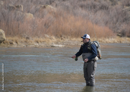 A single man fly fishing on a river with a hat and backpack.