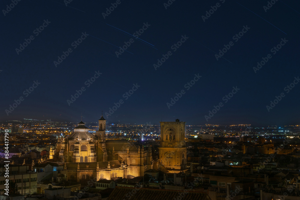 Granada, Spain; August 25 - 2021: The Cathedral of Granada is illuminated in the foreground, shooting stars decorate the night sky and the city lights create a magical atmosphere in the background.