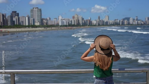 Fashion girl holding hat enjoying view of skyscrapers skyline and blue waving ocean from a promenade terrace. Blurred background.