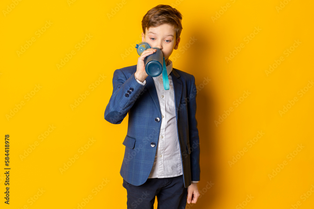 A little boy in a blue suit, white shirt,neatly dressed, drinking water from a bottle. Isolated on yellow. A little boy in a blue suit, white shirt,neatly dressed, drinking water from a bottle. Isolated on yellow.