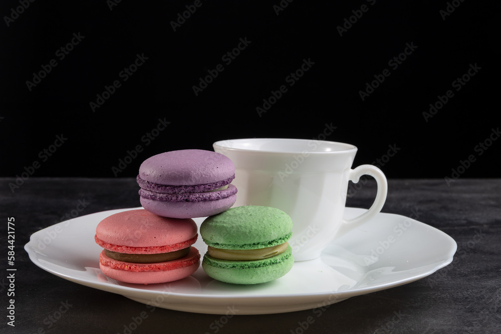 There are macarons on the table in a plate. Behind is a white cup. On a black background. Copy space. There are macarons on the table in a plate. Behind is a white cup. On a black background. Copy space.